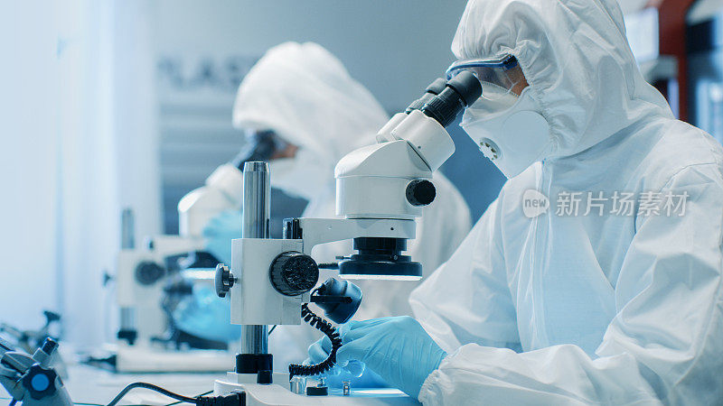 两名工程师/科学家/技术员穿着无菌洁净服使用显微镜进行组件调整和研究。他们在一家电子元件制造厂工作。